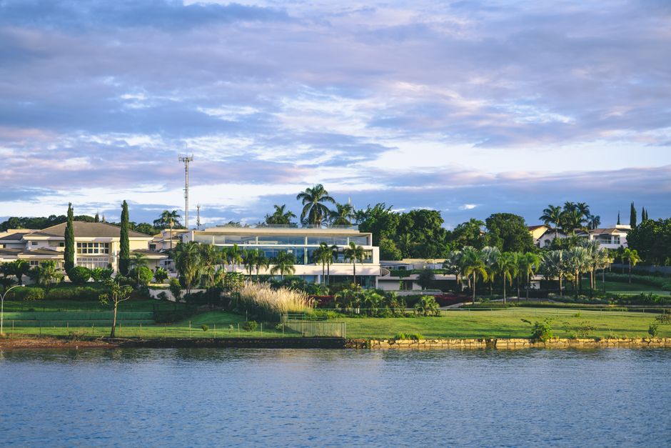 A scenic lakeside property featuring a neatly maintained lawn and garden beds with lush green grass and various plants, including palm trees and shrubs, along with a paved pathway running through the 