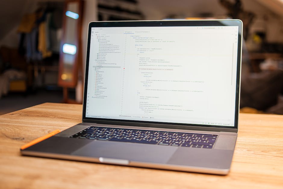 A close-up view of a silver laptop computer on a wooden desk displaying lines of code on its dark screen, with a white smartphone placed nearby, partially out of focus. The scene suggests a workspace 