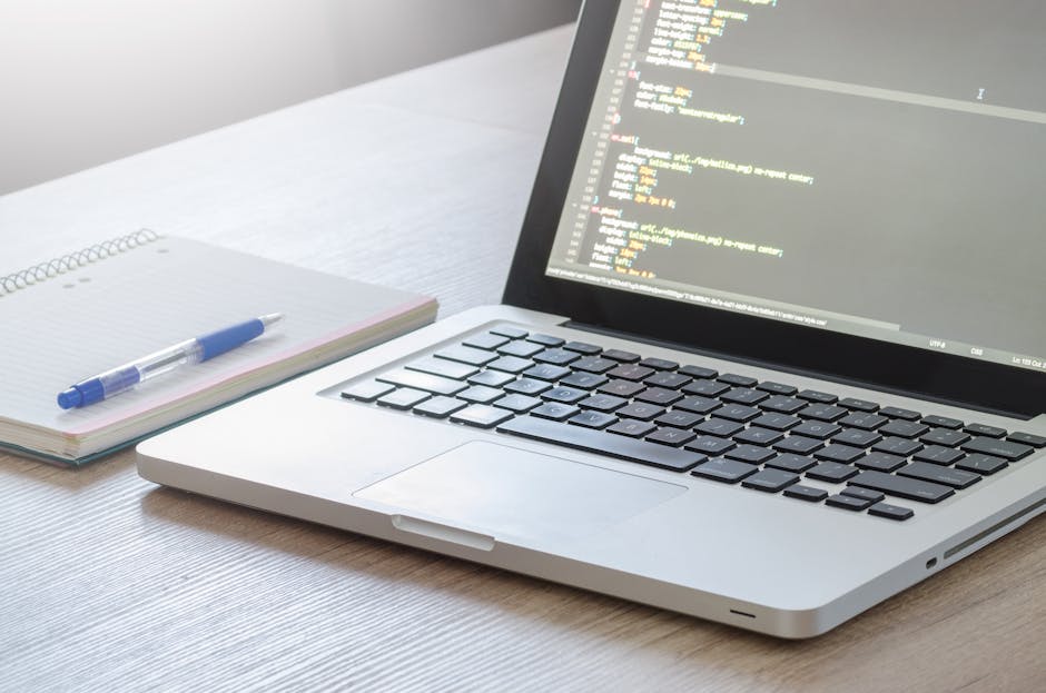 A close-up view of a laptop on a wooden desk displaying lines of code with a backlit keyboard. To the left of the laptop, there is a closed spiral notebook with a blue pen resting on top. The scene is