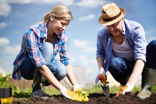 A young woman and an older woman are engaged in gardening outdoors, planting or tending to a garden bed. They are both wearing casual clothing suitable for outdoor work, with the younger woman in a checked shirt and jeans, and the older woman in a light blue shirt, jeans, and a straw hat. They are kneeling on the soil, using small garden trowels to carefully work in the earth. The garden features dark, well-tilled soil, with small seedlings or plants visible in the foreground, and a clear, blue sky overhead, indicating a sunny, pleasant day. The background shows additional garden beds or landscaped areas, suggesting a well-maintained outdoor space designed for gardening activities, which aligns with professional gardening and landscaping services typical of Hainault. This scene exemplifies casual, productive outdoor gardening, highlighting tools, plant care, and the natural environment of a suburban or urban garden in the UK.